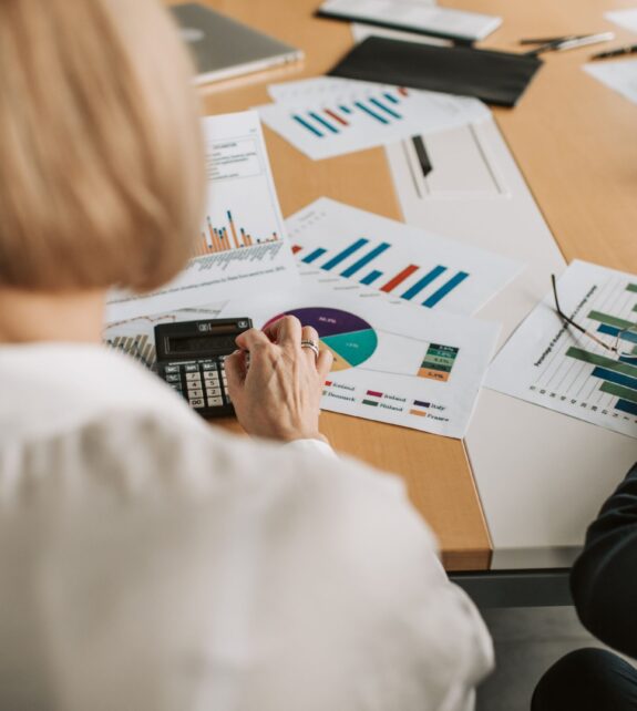 Overhead view of professionals discussing financial graphs and using calculators in an office setting. Blonde woman in a white blouse facing away from the camera is in the foreground.